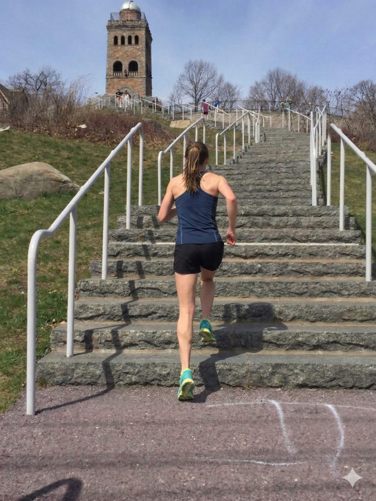 A view looking up the granite stairs of High Rock Tower, Lynn, MA, with a woman runner in motion ascending the steps with other small figures at the summit against a blue sky.